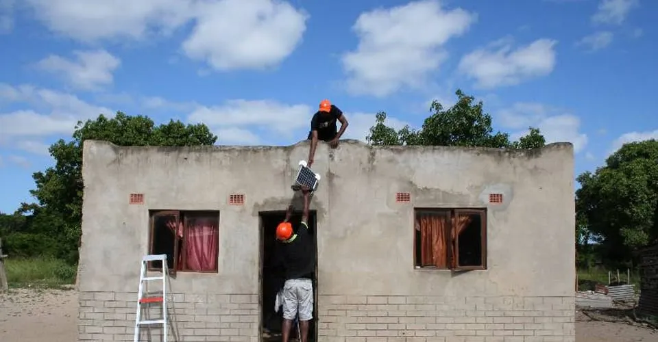 Zwei KINGO Techniker montieren ein Solarbatterie-Panel auf einem Hausdach in ländlichem Afrika