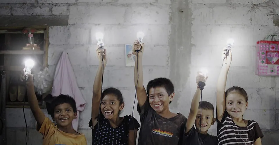 Fünf lachende Kinder halten leuchtende KINGO Solarlampen in einem Betonraum hoch